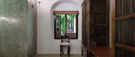 A room with a wooden chair, a bench, a wall-mounted light fixture, and a window with a view of greenery.