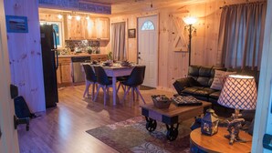 A cozy living room with a dining area, a kitchen in the background, and a wooden exterior wall.
