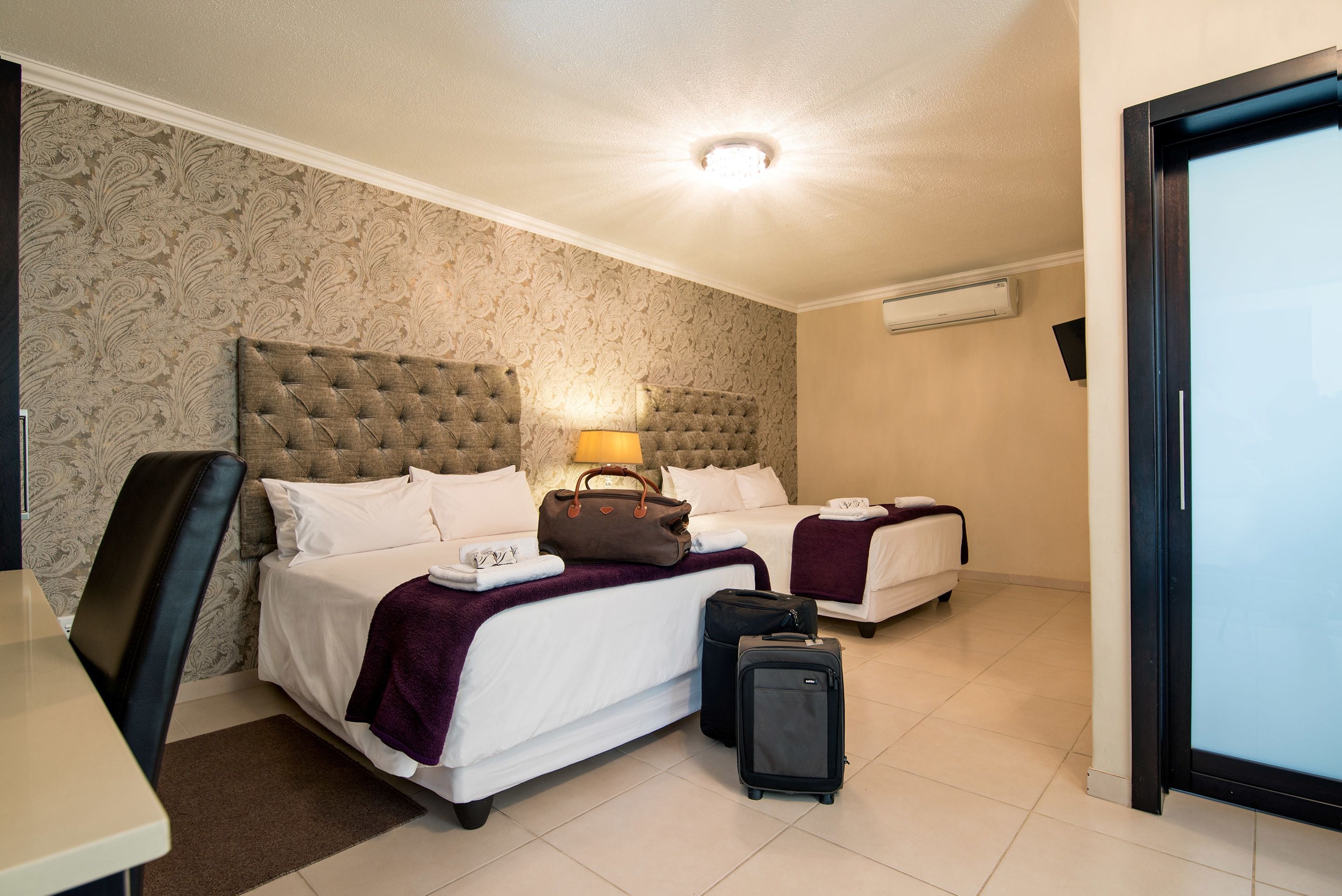 A hotel room with two beds, a desk, and a chair. There is a suitcase and a handbag on the bed.