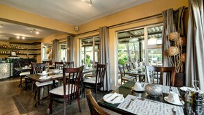 A restaurant with multiple dining tables and chairs, a bar area with bottles, and large windows allowing natural light.