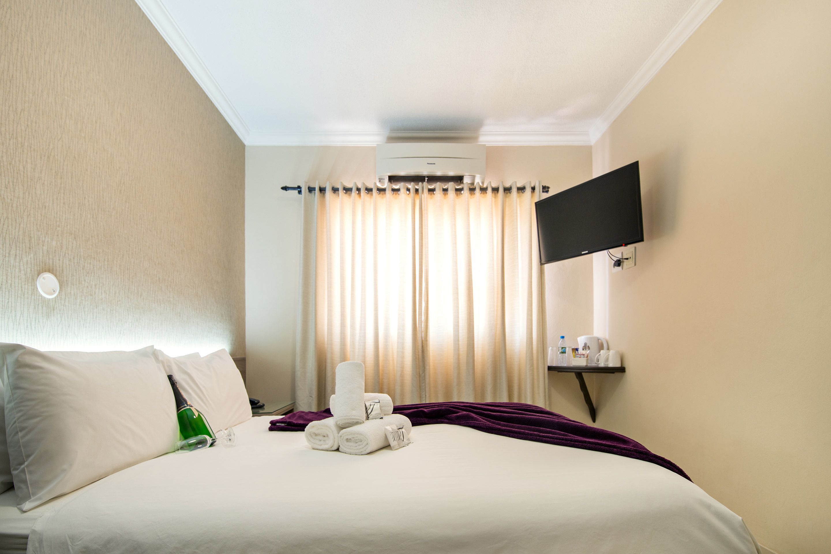 A hotel room with a bed, a flat-screen TV mounted on the wall, a window with curtains, and a small table with toiletries.