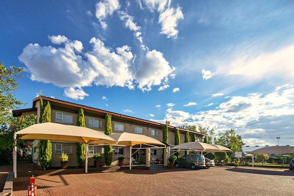 A hotel with a brick facade, outdoor seating with umbrellas, and a parking area. - Auas City Hotel (Windhoek)