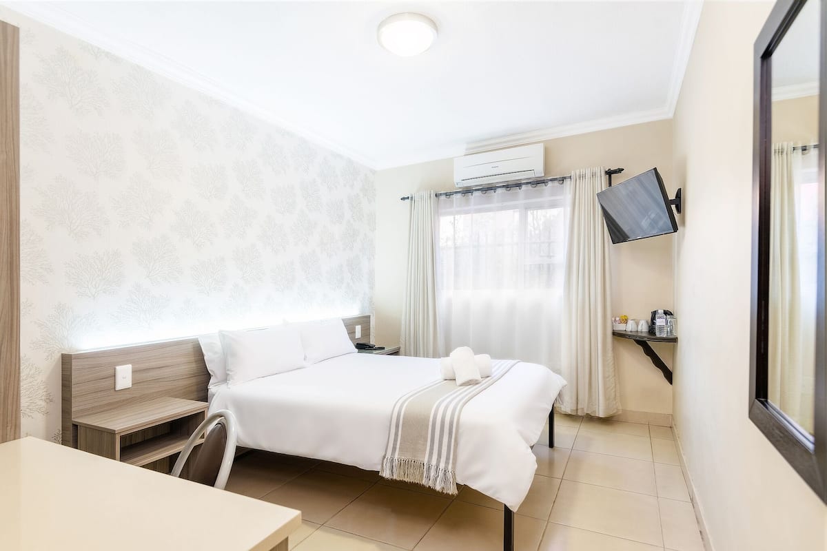 Una habitación de hotel con cama, mesita de noche, silla, televisor y ventana con cortinas.