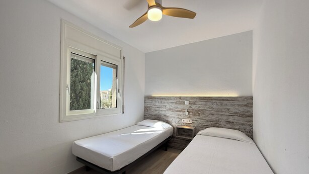 A bedroom with a bed, a bench, a ceiling fan, a window, and a wall-mounted light fixture. - Agi Rescator 212 Apartamento for rent in Roses Girona. (Rosas / Roses)