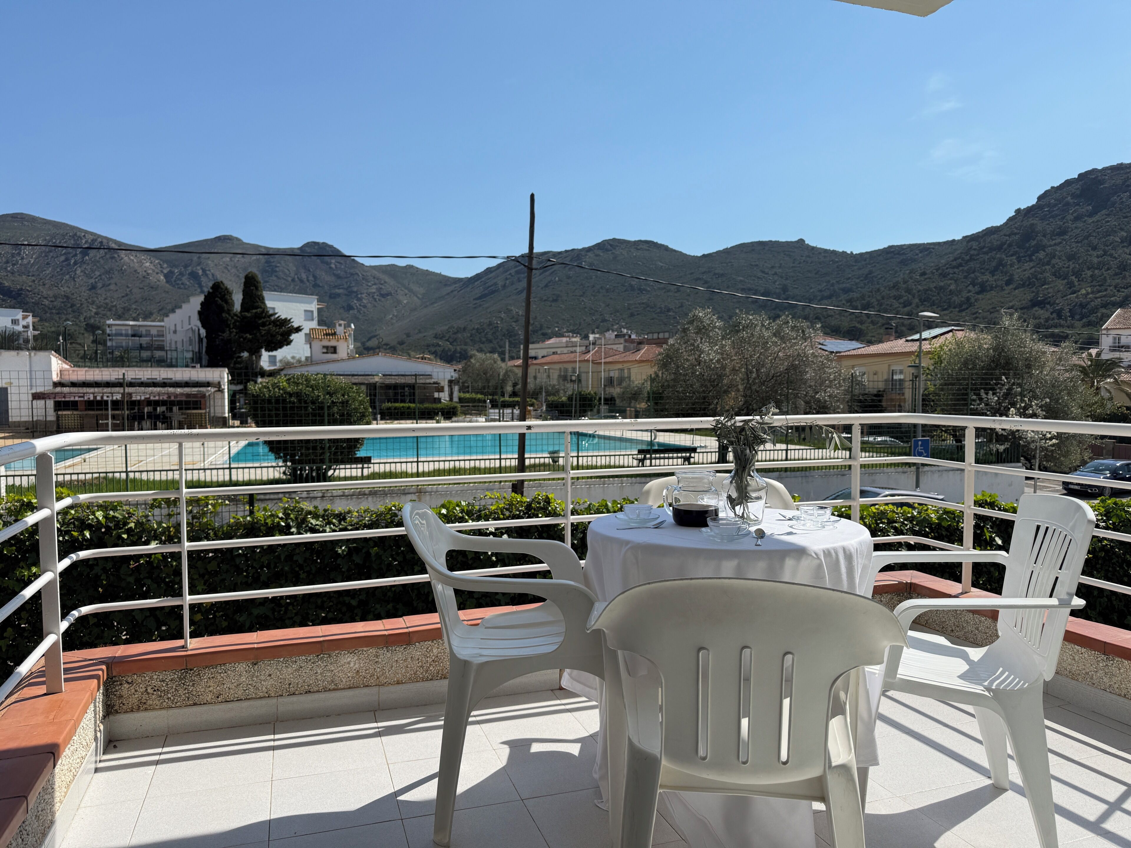 A terrace with a table and chairs, overlooking a swimming pool and mountains.