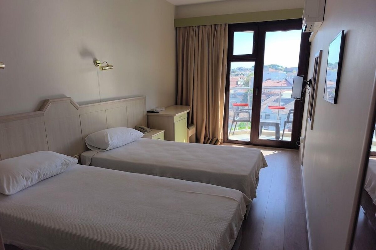 Une chambre d’hôtel avec deux lits, un bureau, une chaise, une fenêtre avec des rideaux et une vue sur les bâtiments environnants.