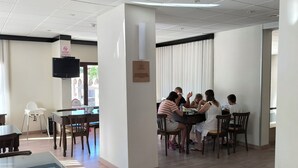 A dining area with wooden tables and chairs, a flat-screen TV mounted on the wall, and a ceiling with a grid pattern. - Ridvan Hotel (Cesme)