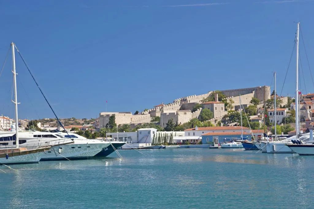 Un puerto deportivo con barcos amarrados, una estructura que parece un castillo al fondo y un cielo despejado.