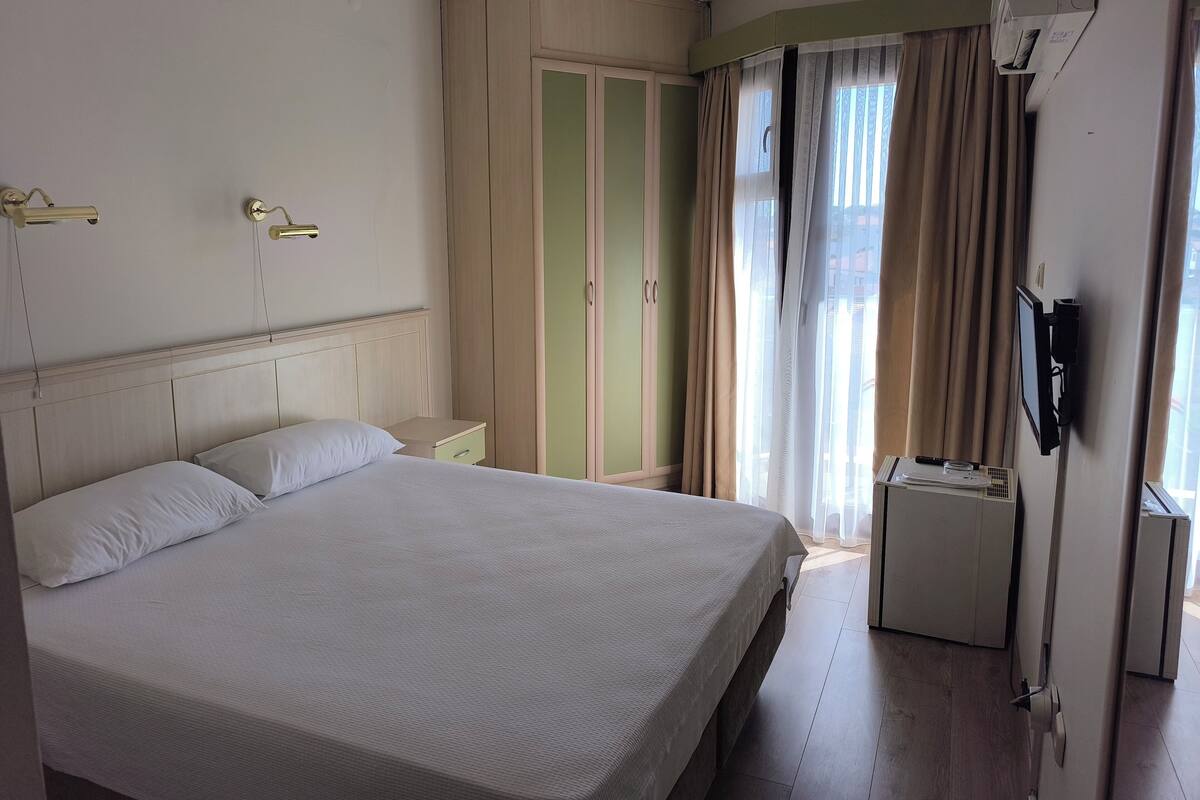 Habitación de hotel con cama, mesitas de noche, armario, televisor y ventana con cortinas.