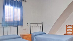 A bedroom with two single beds, a wooden nightstand, a metal headboard, a wooden chair, and a window with blue curtains.
