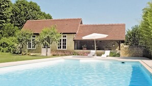 Una piscina con un paraguas blanco, una casa con techo marrón y un muro de piedra.