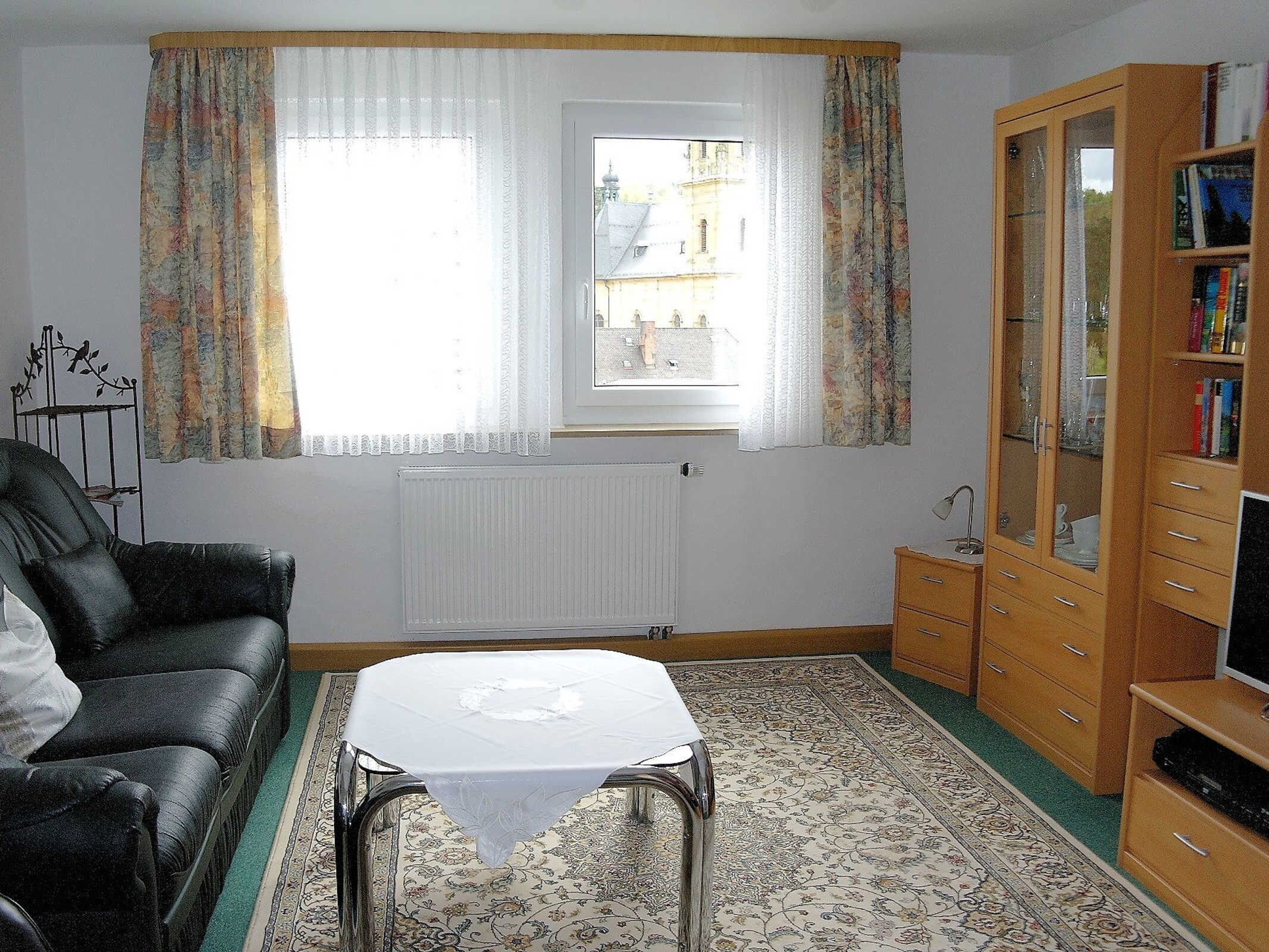 Ein Wohnzimmer mit einem schwarzen Ledersofa, einem kleinen Tisch, einem Heizkörper, einem Fenster mit Vorhängen und einem Holzschrank mit Glastüren.