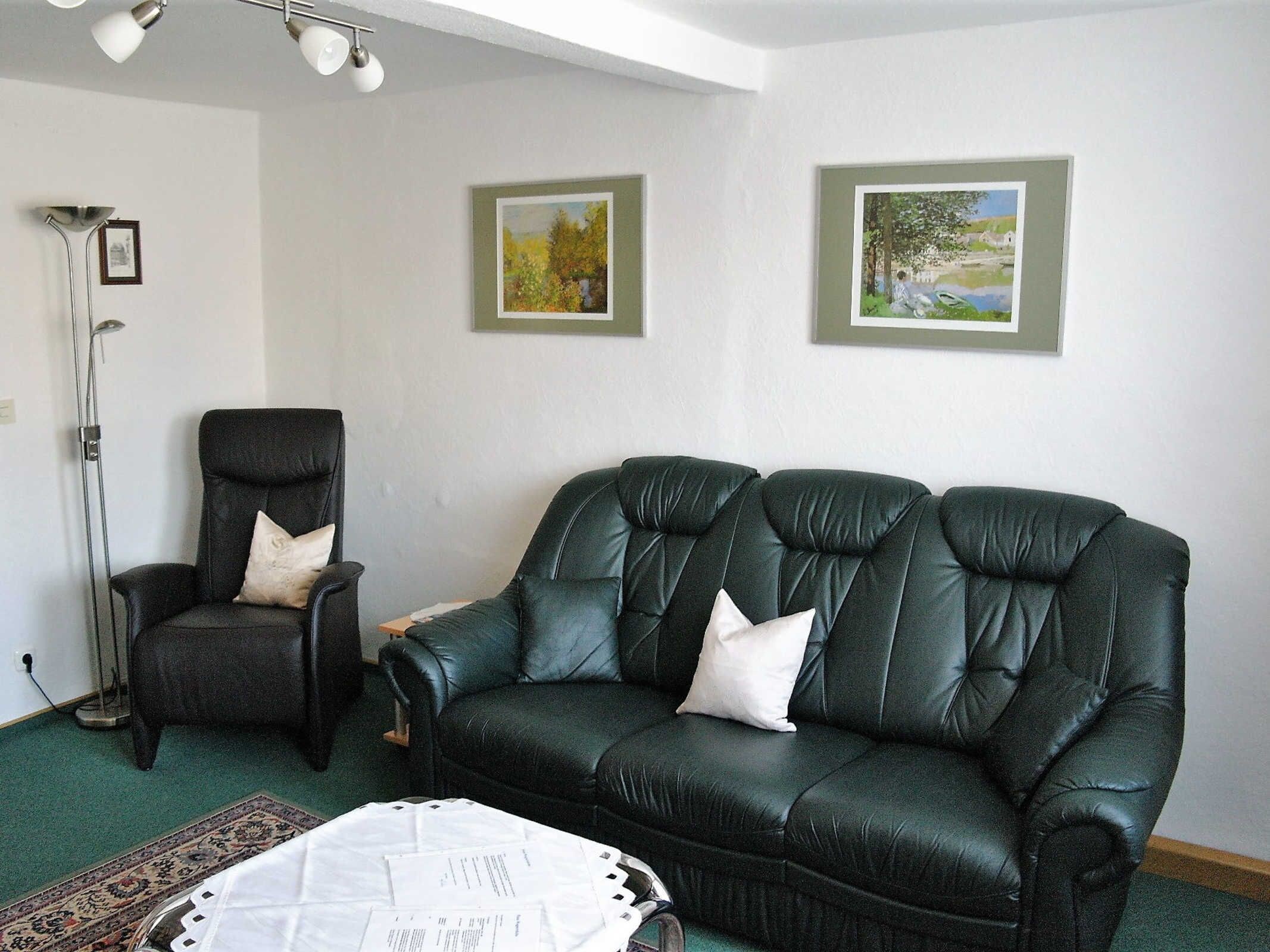 A living room with a dark leather sofa, a black armchair, and a floor lamp. There are two framed paintings on the wall.