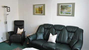 A living room with a dark leather sofa, a black armchair, and a floor lamp. There are two framed paintings on the wall.