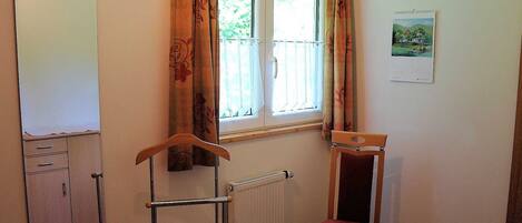 Une pièce avec un radiateur, une chaise en bois dotée d’un coussin rouge, un tableau blanc accroché au mur et une fenêtre avec des rideaux.