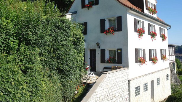 Ein zweistöckiges weißes Gebäude mit rotem Ziegeldach, zahlreichen Fenstern mit Fensterläden und Blumenkästen im oberen Geschoss.