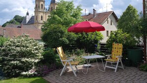 Eine Terrasse mit zwei weißen Stühlen und einem roten Sonnenschirm, umgeben von üppigem Grün und einem Pflasterweg.