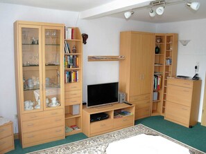 A bedroom with a TV, wooden wardrobe, and shelves with books and decorative items.