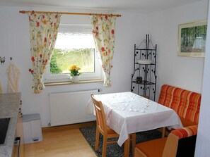 A cozy room with a dining table, a bench, a radiator, a window with curtains, and a shelf with items. - Holiday house Wagnershöhe (Gößweinstein)
