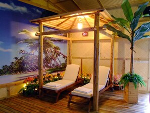 A wooden deck with two lounge chairs under a pergola, a tropical mural, and a potted plant.