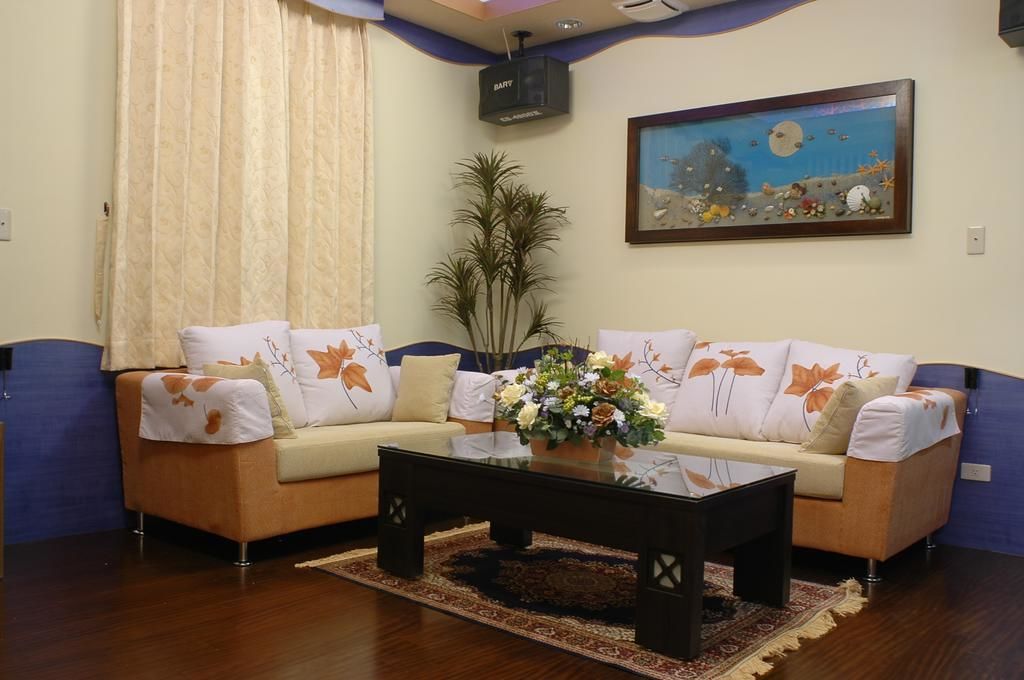 A living room with a beige sofa, white cushions with orange leaf patterns, a glass coffee table with a floral centerpiece, a tall plant, and a painting of a beach scene.