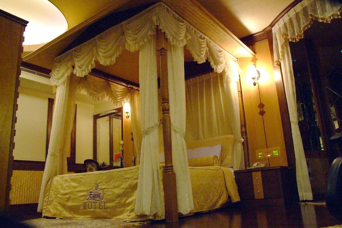 Un lit à baldaquin avec quatre poteaux, surmonté d’une couvre-lit matelassé, une table de chevet, une lampe et un miroir, dans une chambre d’hôtel.