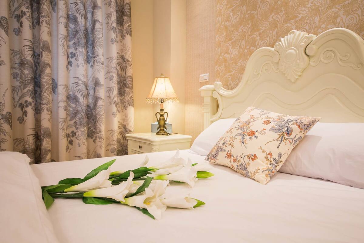 A neatly made bed with white linens, a floral pillow, and a bouquet of white flowers on the bed. There is a nightstand with a lamp and a small table beside the bed.