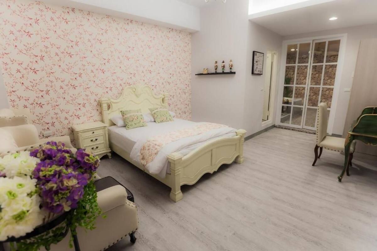 A bedroom with a floral wallpaper, a bed with a decorative headboard, a bedside table, a chair, and a window with a view.