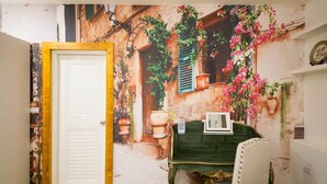 A room with a white door, a green table, and a chair. There is a mural on the wall depicting a street scene with buildings and plants. - England Garden (Tainan)