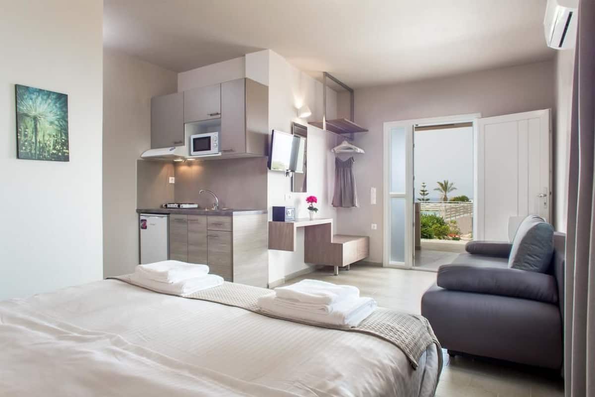 A modern hotel room with a bed, a sofa, a kitchenette, and a balcony with plants.