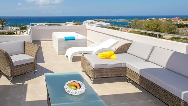 A rooftop terrace with wicker furniture, a glass table with fruit, and a view of the sea.