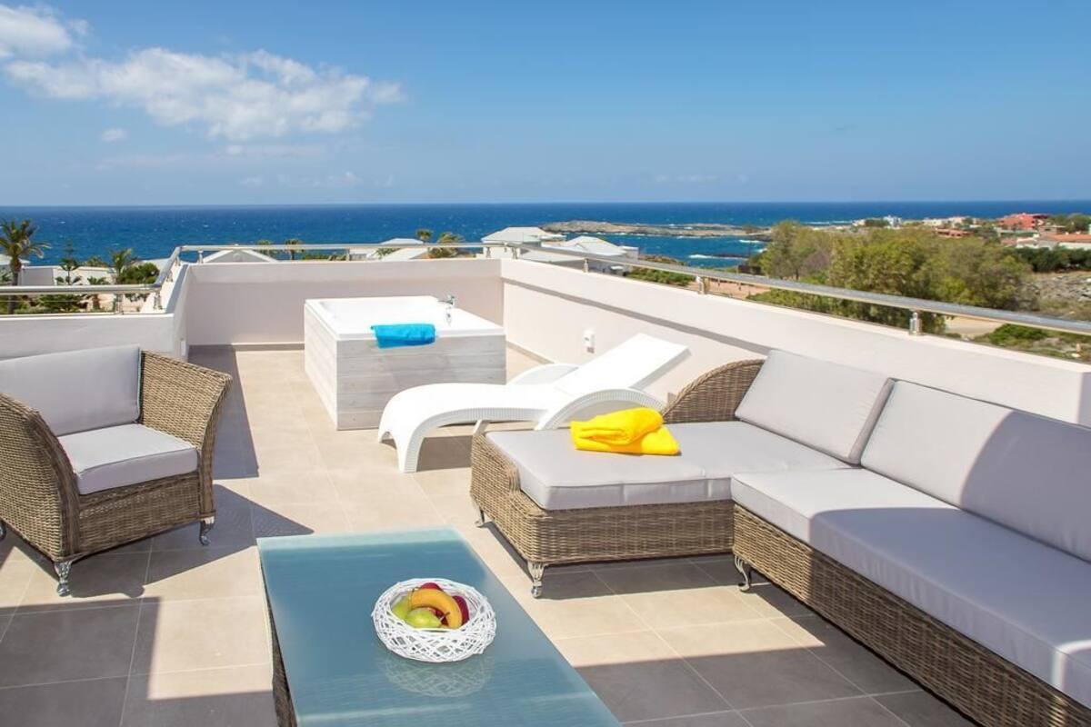 A rooftop terrace with wicker furniture, a glass table with fruit, and a view of the sea.