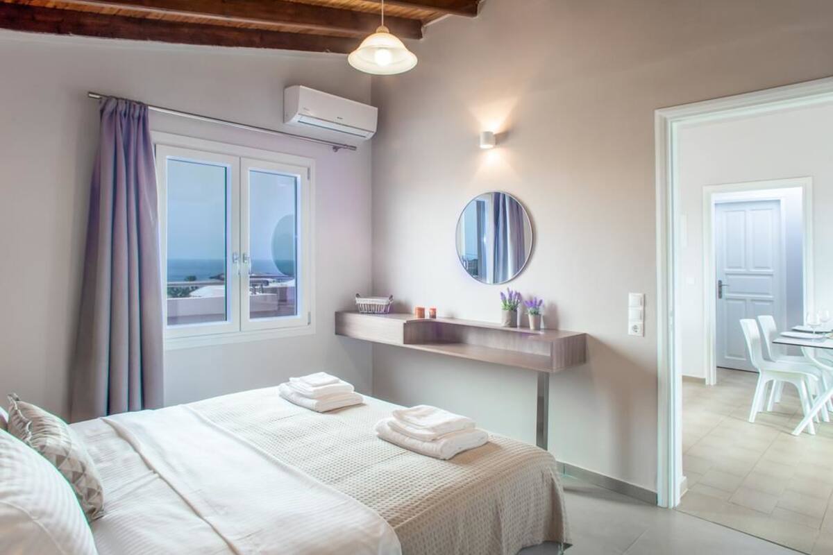 A bedroom with a bed, a window with curtains, a bedside table with toiletries, and a view of the outside.