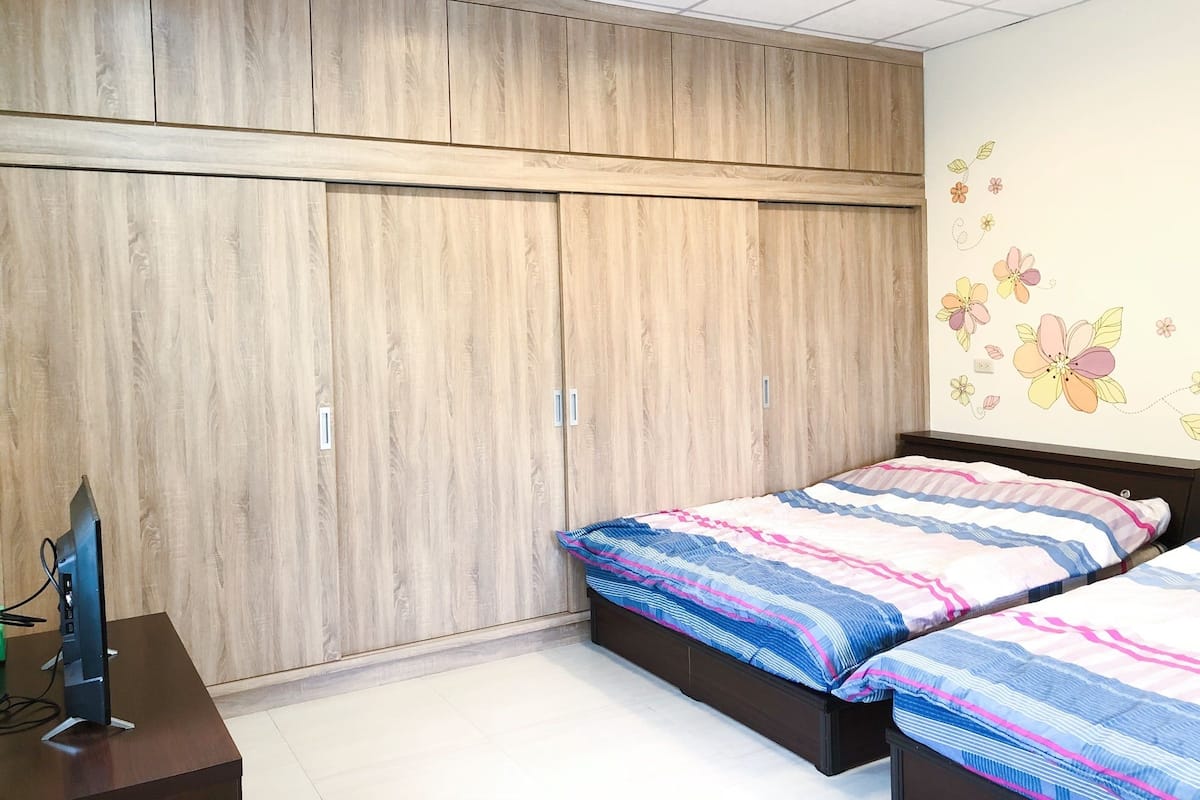 A bedroom with two beds, a wooden wardrobe, and a TV on a desk.