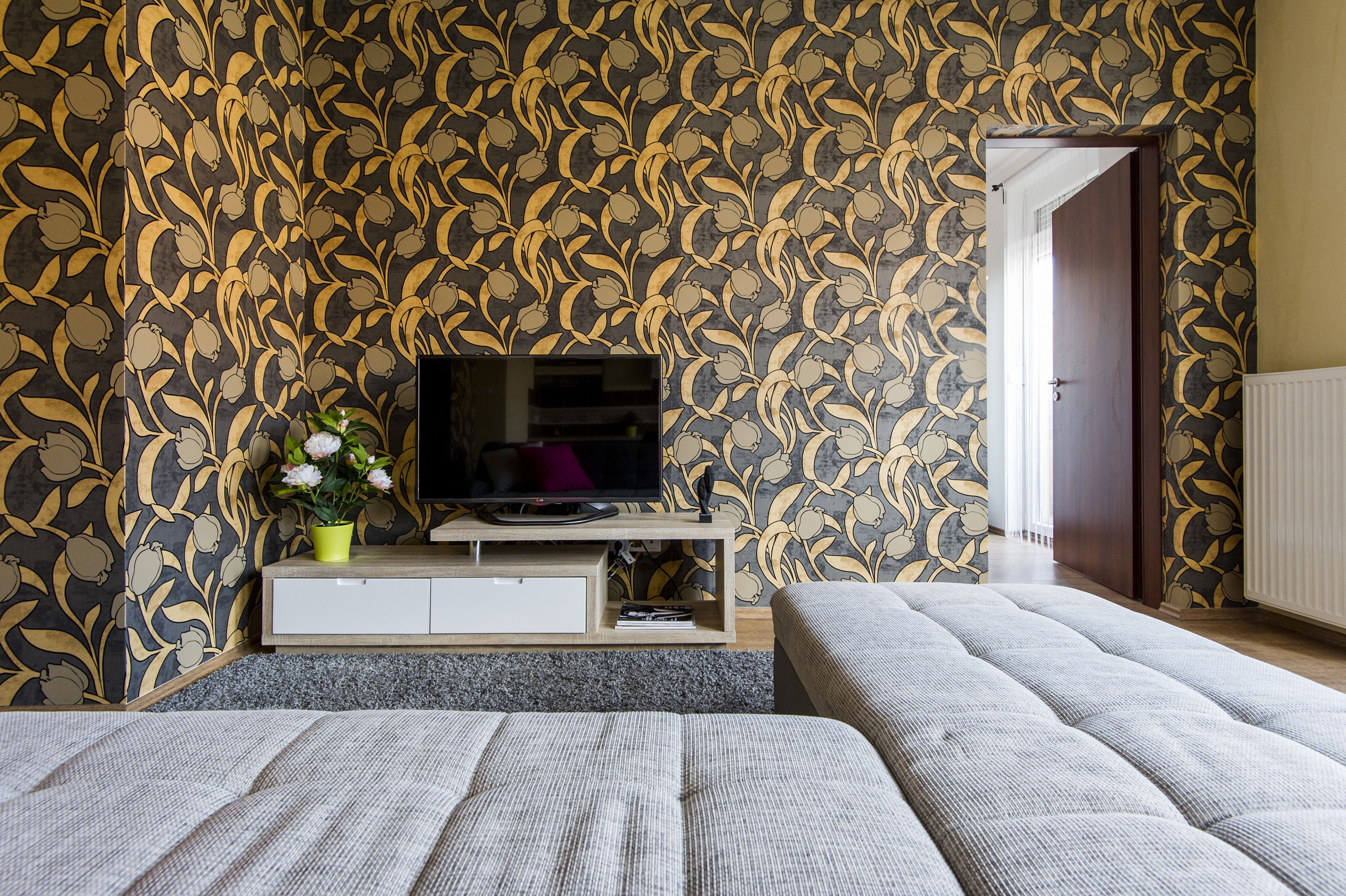 Ein Schlafzimmer mit Fernseher auf einem Holzbalken, einer Topfpflanze und einem Bett mit grauer Decke.