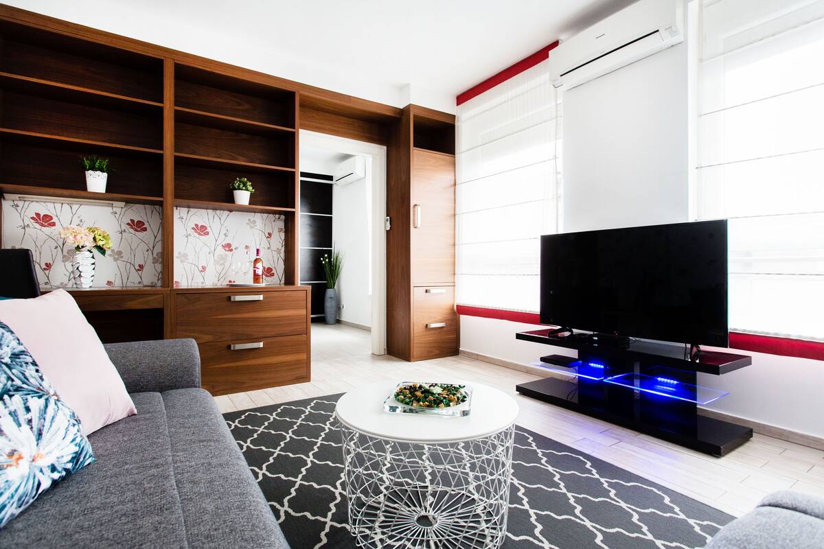 A modern living room with a grey sofa, a coffee table, a TV on a stand, and a wooden shelving unit with plants.