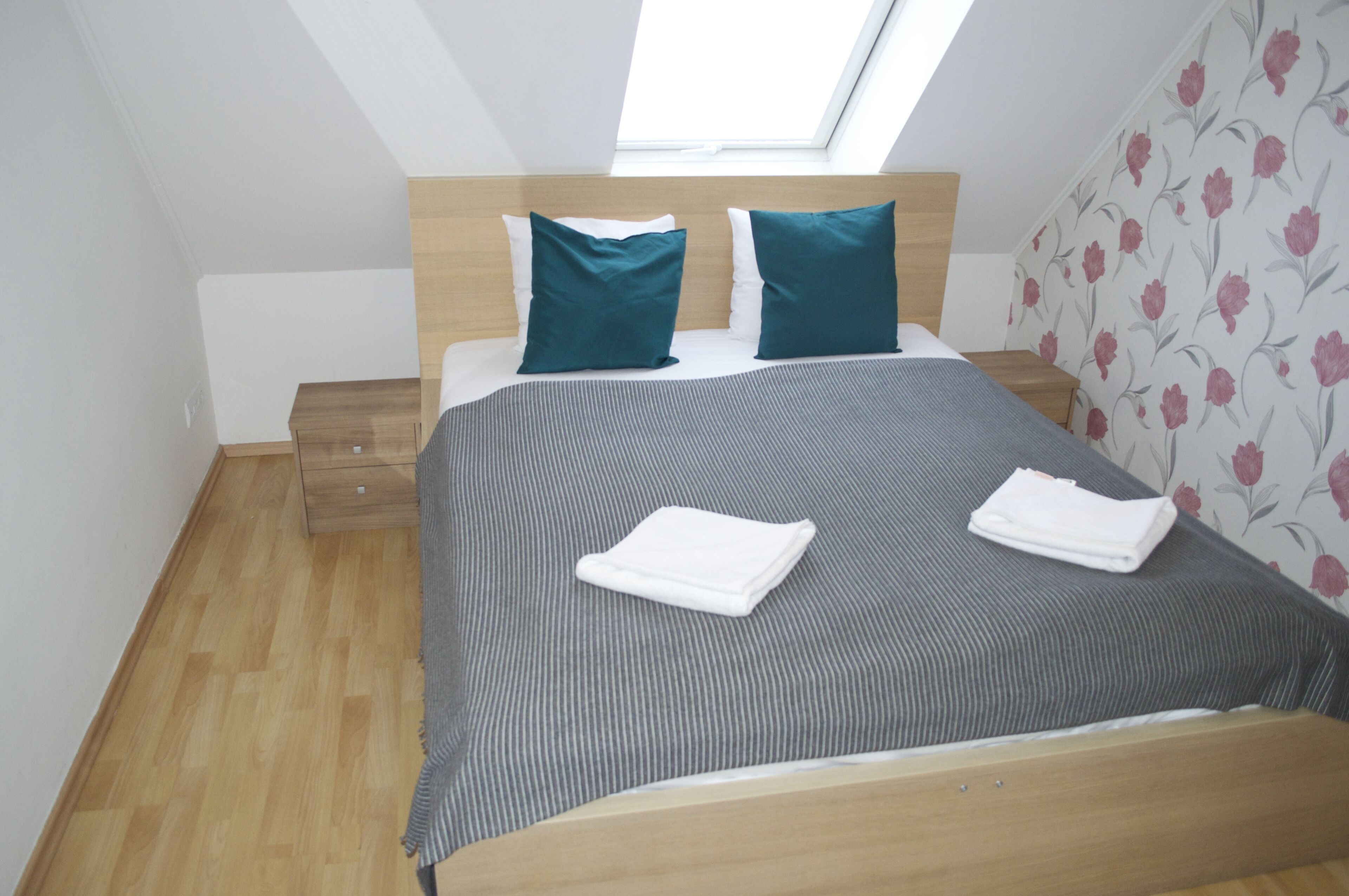 Ein ordentlich gemachtes Bett mit gestreifter Bettdecke, zwei Kissen und gefalteten Handtüchern. Ein Nachttisch aus Holz mit Schublade, ein Fenster mit Blick auf den Himmel und Blumenwallpaper an der Wand sind ebenfalls zu sehen.