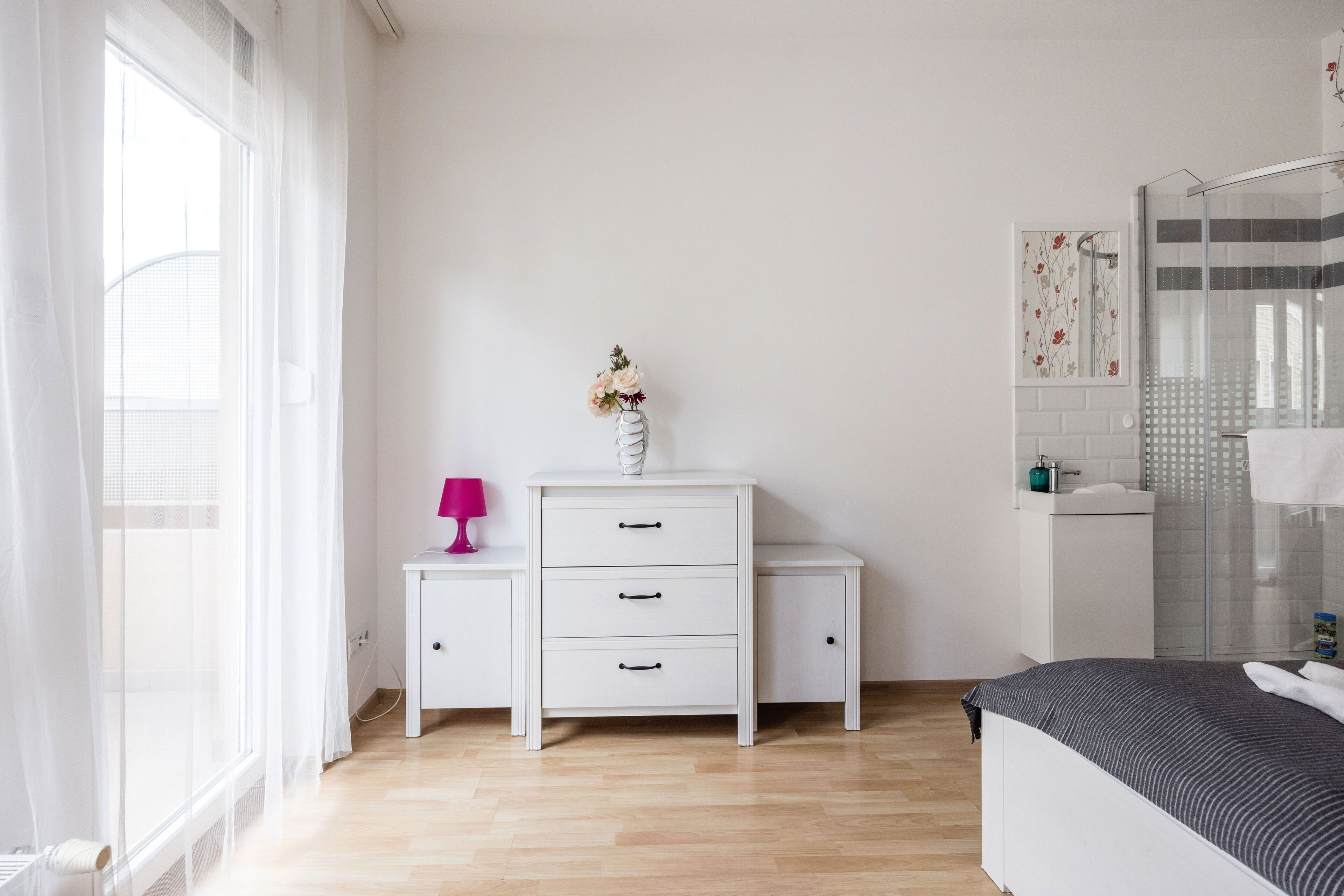 A bedroom with a bed, a bedside table, a dresser, a window with sheer curtains, and a shower area with a glass door.