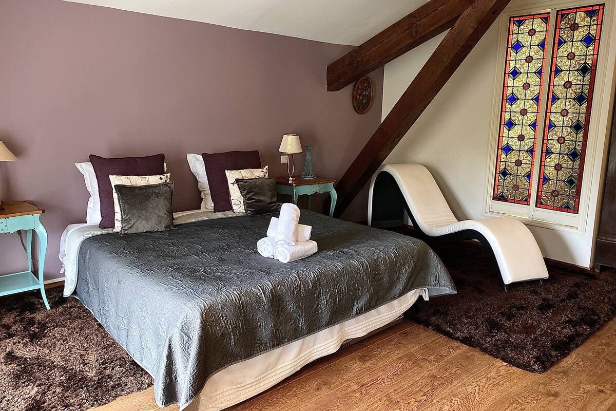 Een slaapkamer met een bed, kussens, een stoel, een lamp en een glas-in-loodraam.