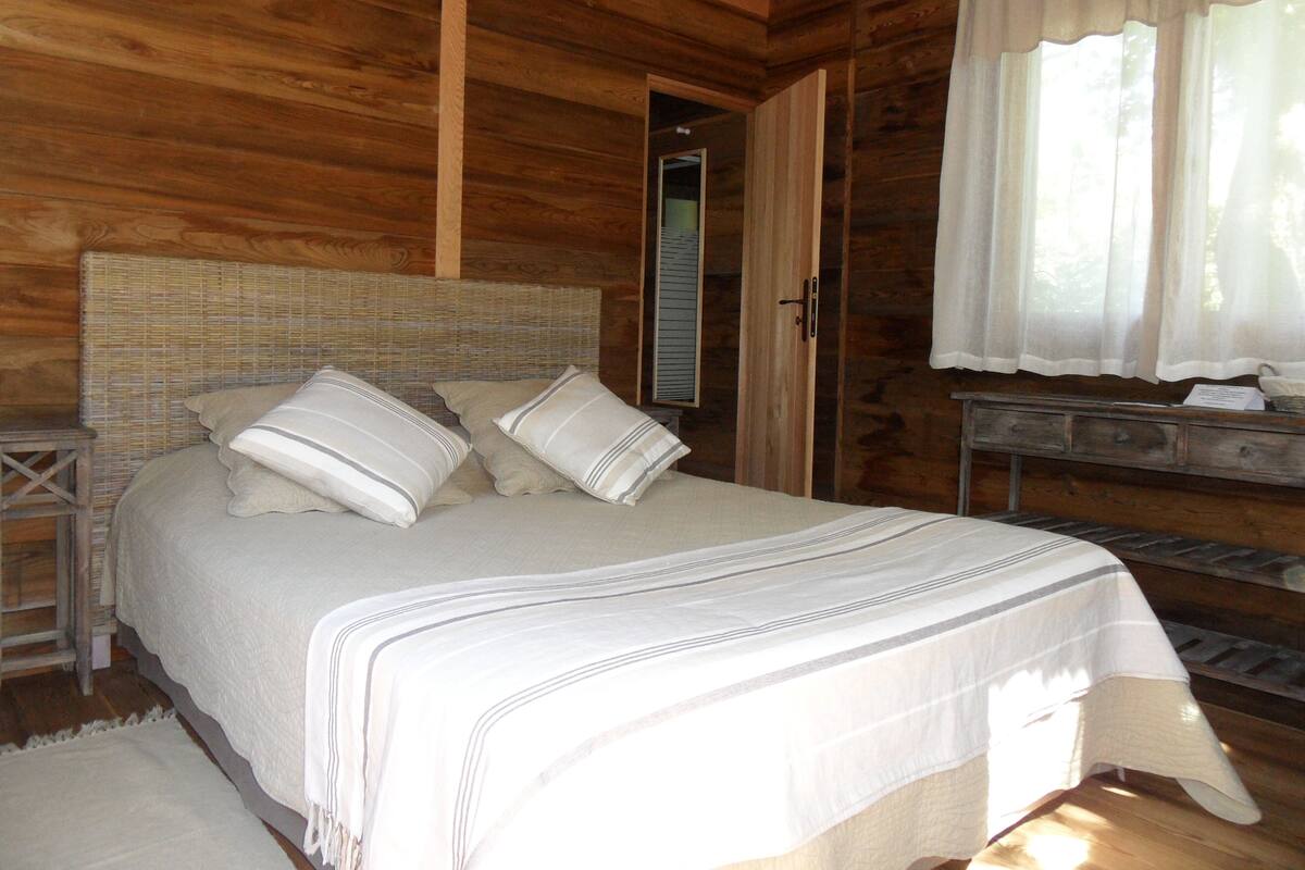 Een houten slaapkamer met een bed, kussens, een nachtkastje en een raam met gordijnen.