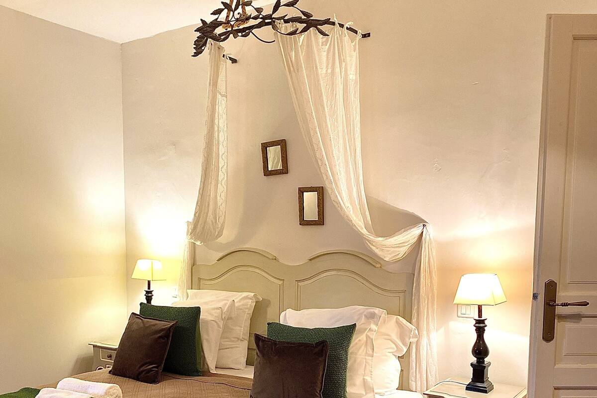 Een net opgemaakt bed met een groene sprei en meerdere kussens, twee nachtkastjes met lampen, en een baldakijnbed.