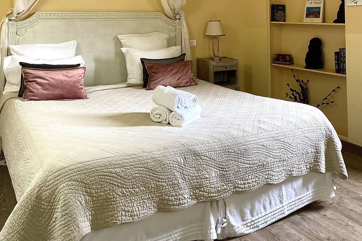 Een net opgemaakt bed met een witte dekbedovertrek en twee kussens, een nachtkastje met een lamp, en een plank met boeken en decoratieve objecten.