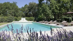 Une piscine rectangulaire avec une fontaine au centre, entourée de chaises longues et d’une pergola dotée d’un parasol.