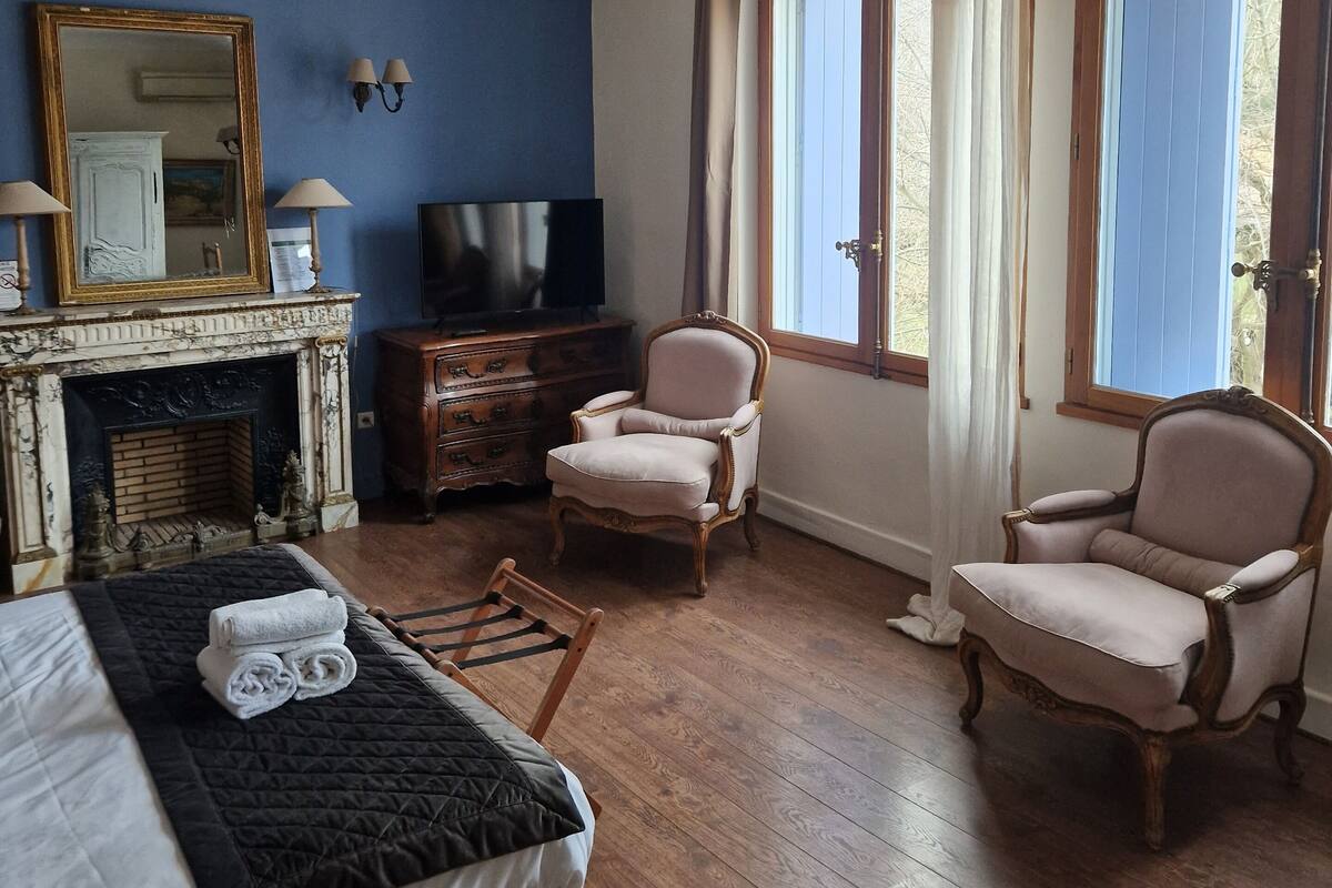Een kamer met een bed, twee fauteuils, een televisie, een open haard en grote ramen.