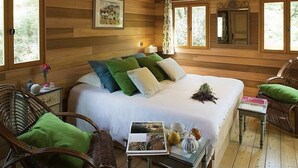 Une pièce de style cabane en bois, avec un grand lit, des fauteuils en osier, une petite table avec un livre et une tasse, et une fenêtre avec des rideaux.