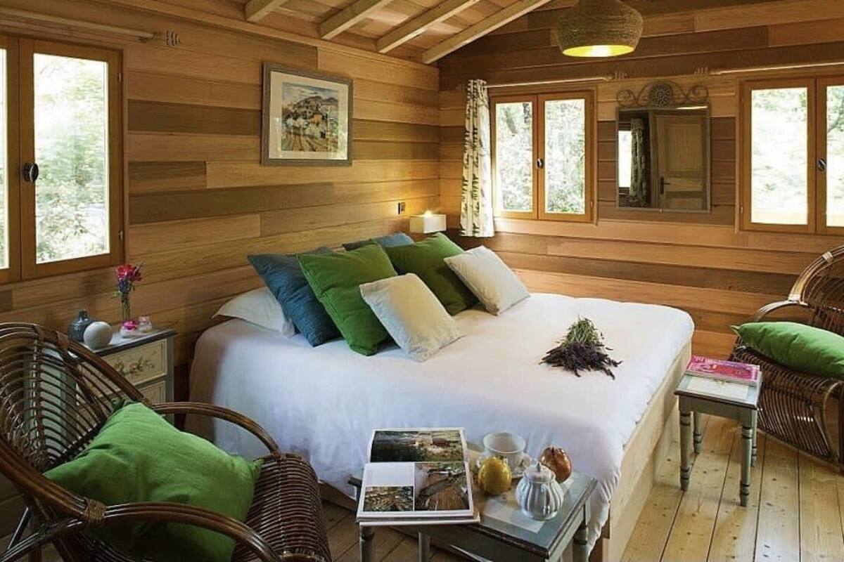 Een houten kamer met een groot bed, rieten stoelen, een klein tafeltje met een boek en een beker, en een raam met gordijnen.