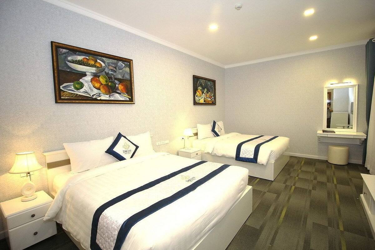 Quarto de hotel com duas camas, mesa de cabeceira com abajur, espelho e obras de arte nas paredes.