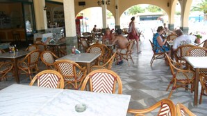 A covered outdoor dining area with wooden chairs and tables, a bar in the background, and a view of the street. - Lemon Grove Hotel and Studios (Corfu)
