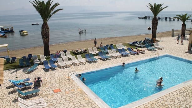 A pool area with lounge chairs and a swimming pool, overlooking a beach with boats and palm trees. - Lemon Grove Hotel and Studios (Corfu)