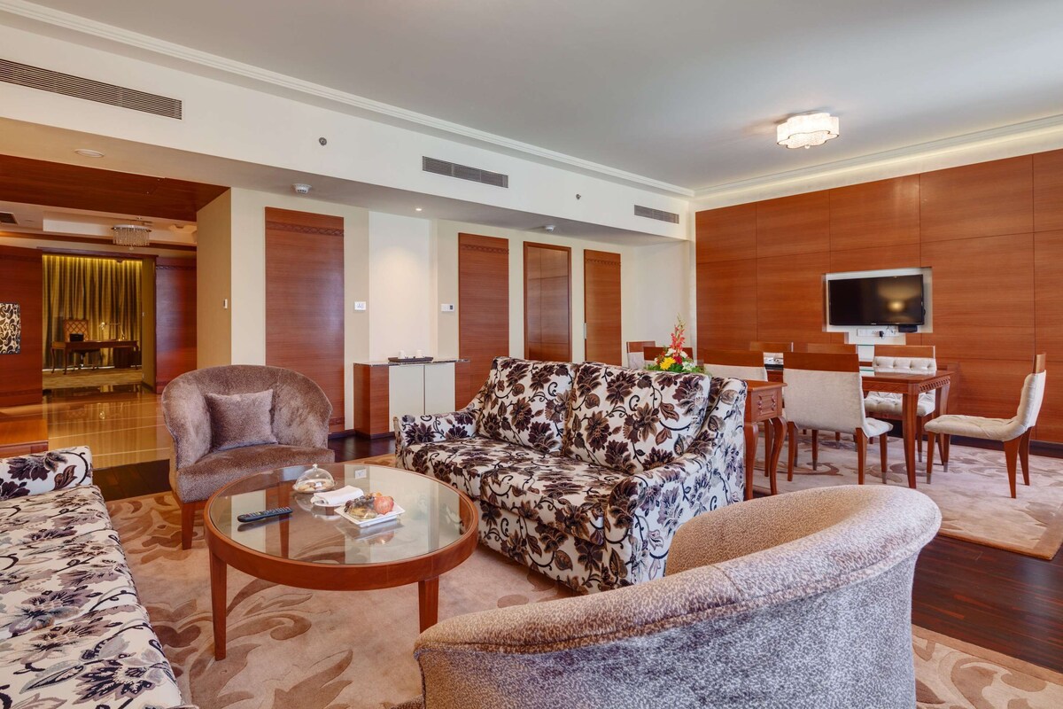 A hotel lobby with a sofa, armchairs, a coffee table, a TV, a dining area, and a reception desk.